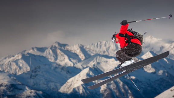 downhill skier in red jacket mid jump with snowy mountains in the background