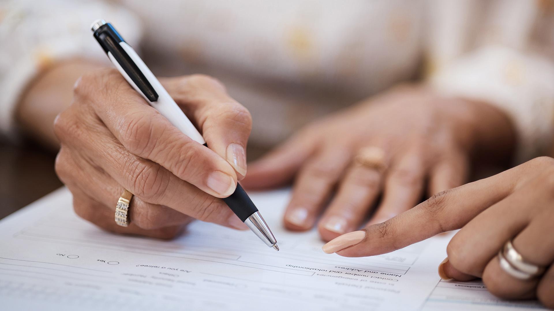 Hands holding a pen and pointing to an area on a legal document to sign.