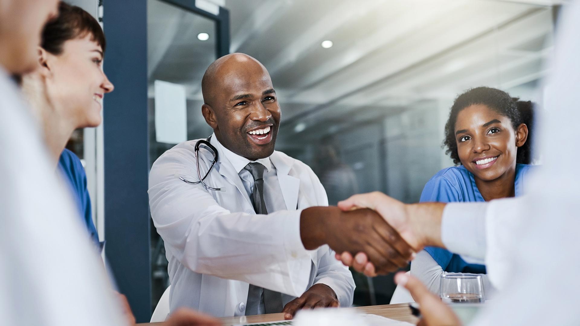 A smiling doctor shakes hands with another healthcare professional, surrounded by smiling colleagues.