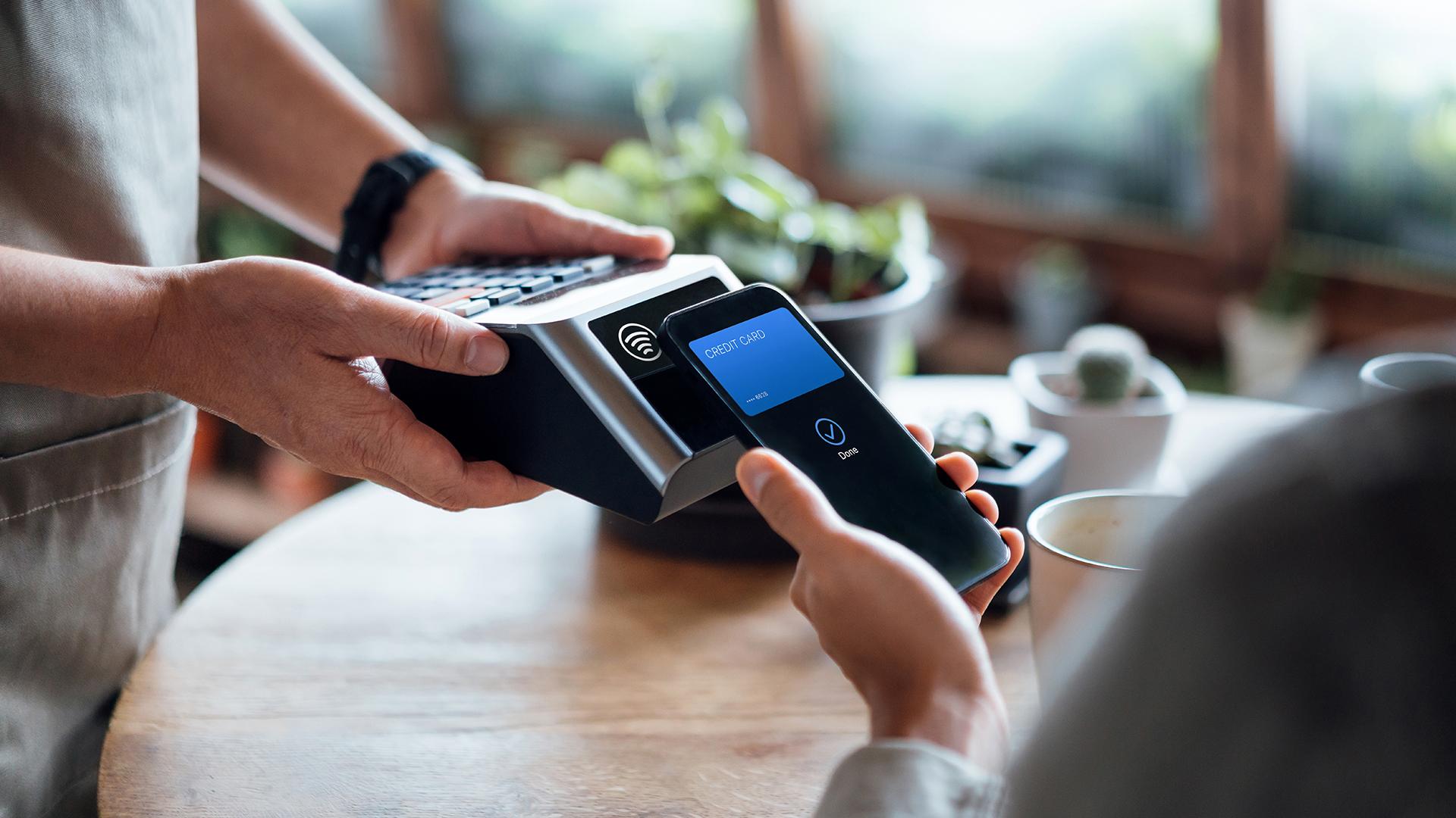 Close up of a male's hand paying bill with credit card contactless payment on smartphone in a cafe, scanning on a card machine