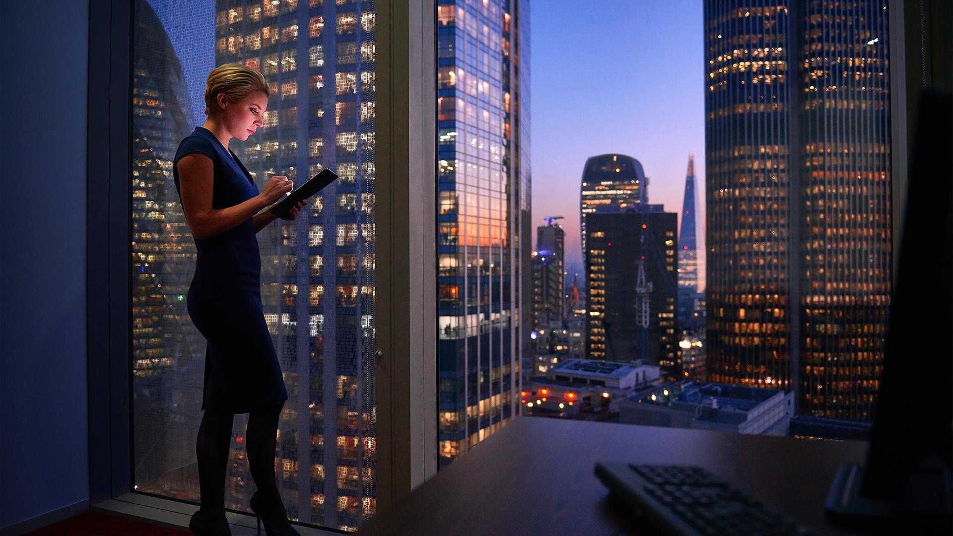 A woman working on her iPad next to a floor to ceiling window overlooking a city skyline.