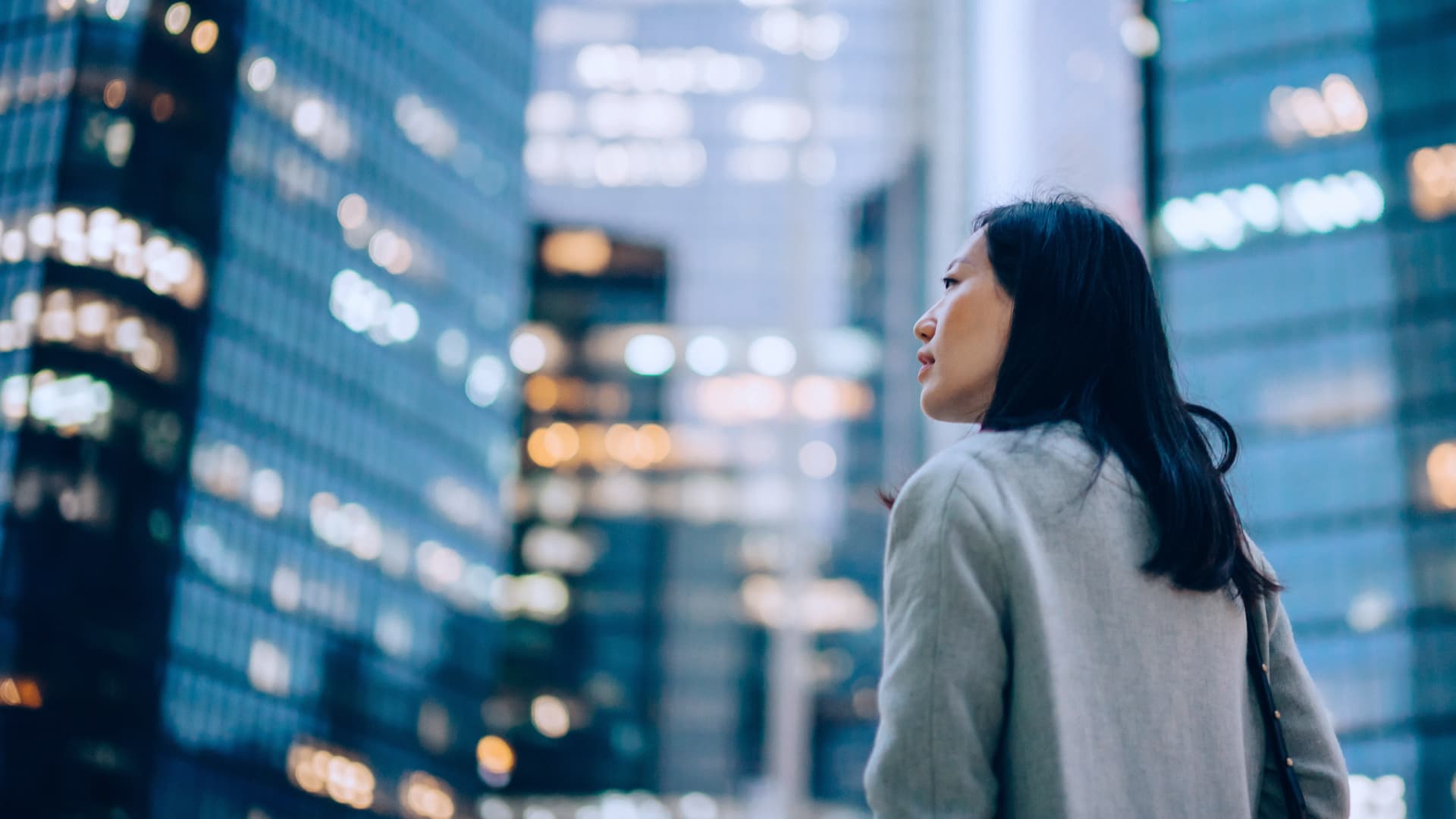 Low angle side profile of confident and professional young Asian businesswoman looking up while standing against contemporary corporate skyscrapers with illuminated facade in financial district in the evening. Female leadership and determined to success