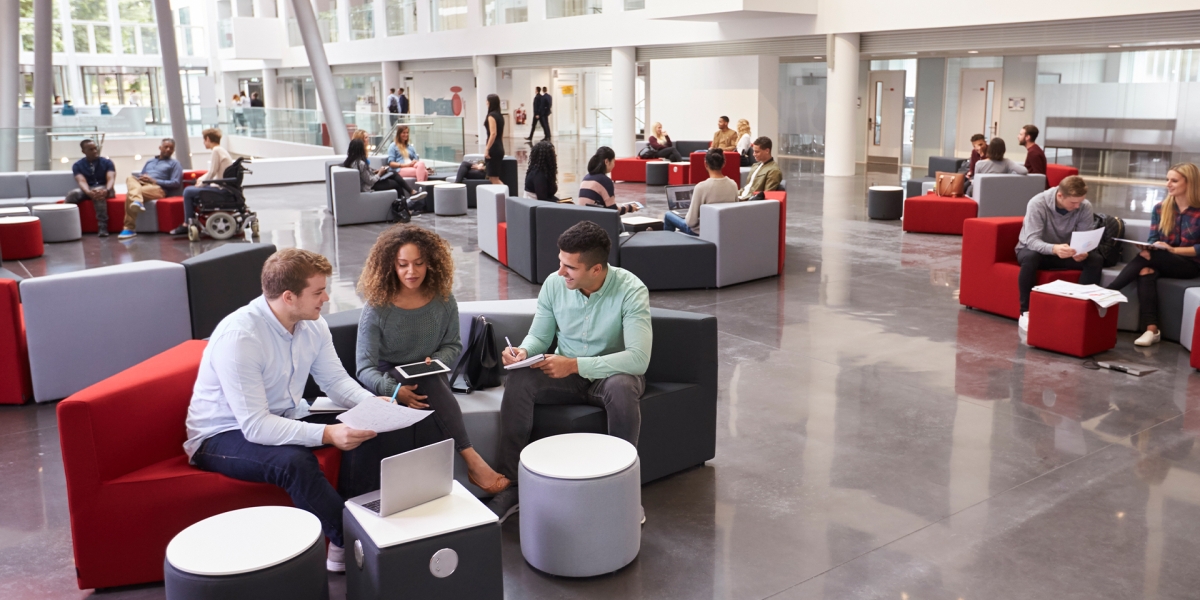 A group of students in a common area of a university building  with red and grey seating 