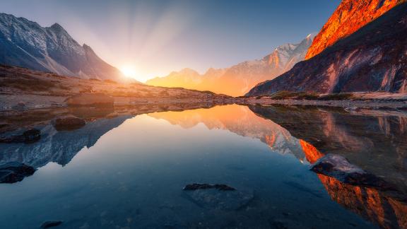 Beautiful landscape with high mountains with illuminated peaks, stones in mountain lake, reflection, blue sky and yellow sunlight in sunrise. Nepal.