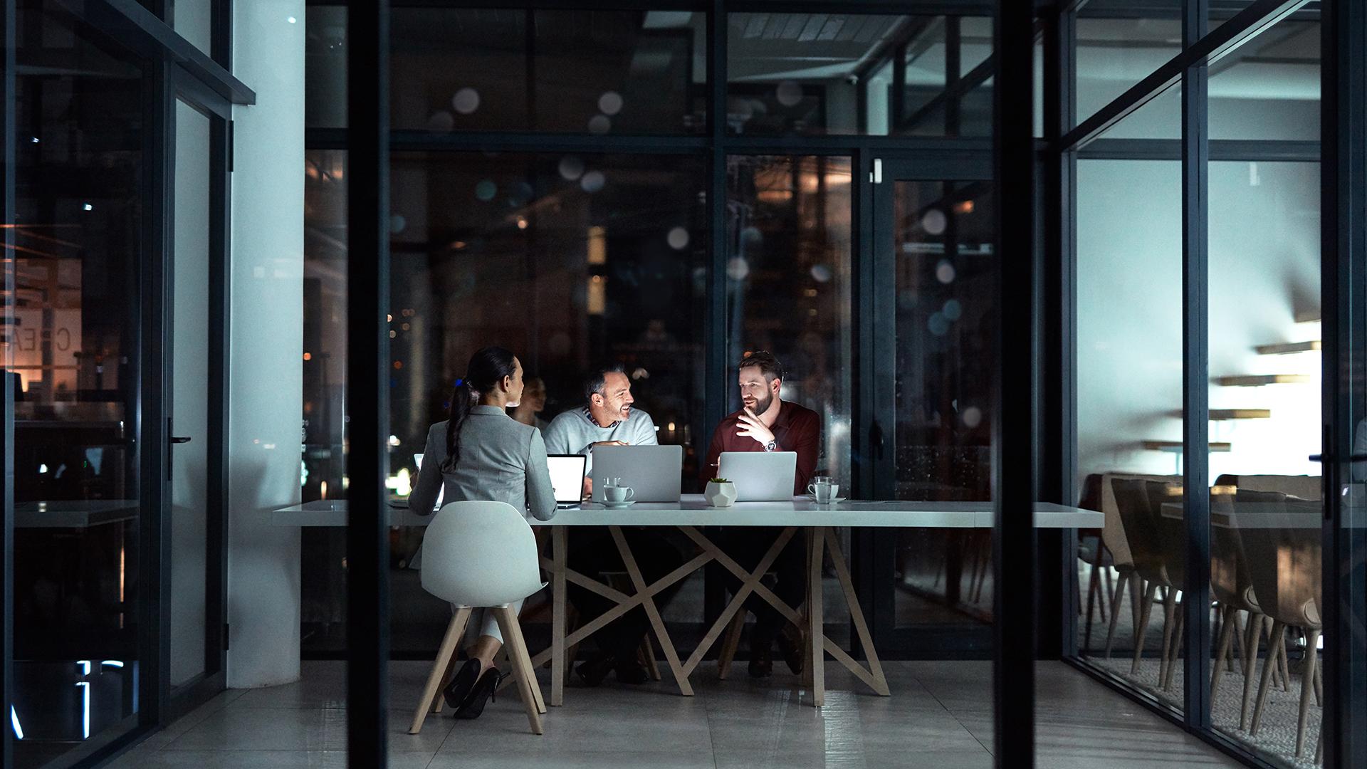 A woman and two male colleagues sit in a dimly lit office at dusk