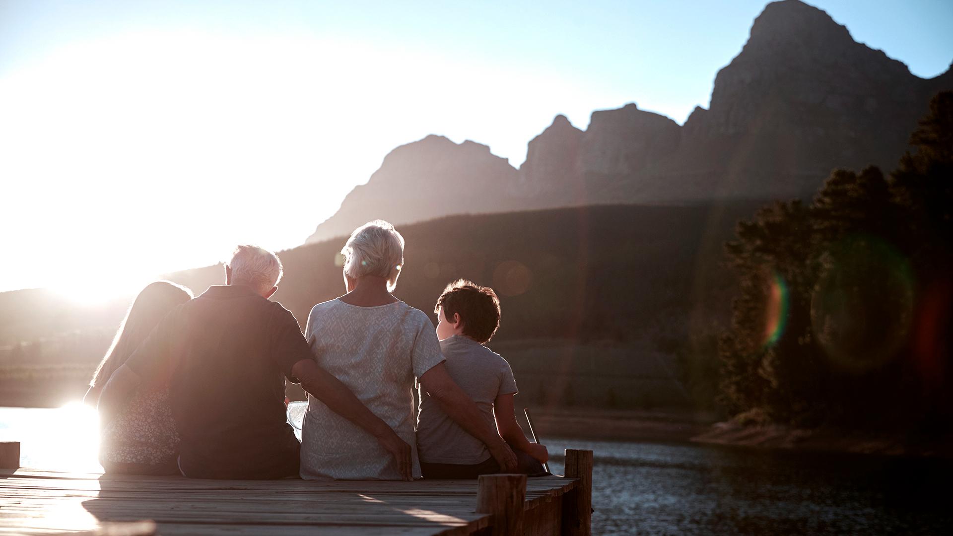 A family of four sits on the edge of a lake dock admiring the sunset behind ocean cliffs.