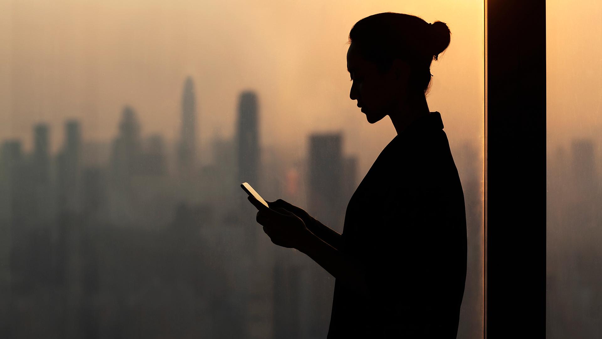 Silhouette of young woman using smartphone next to window with cityscape - stock photo