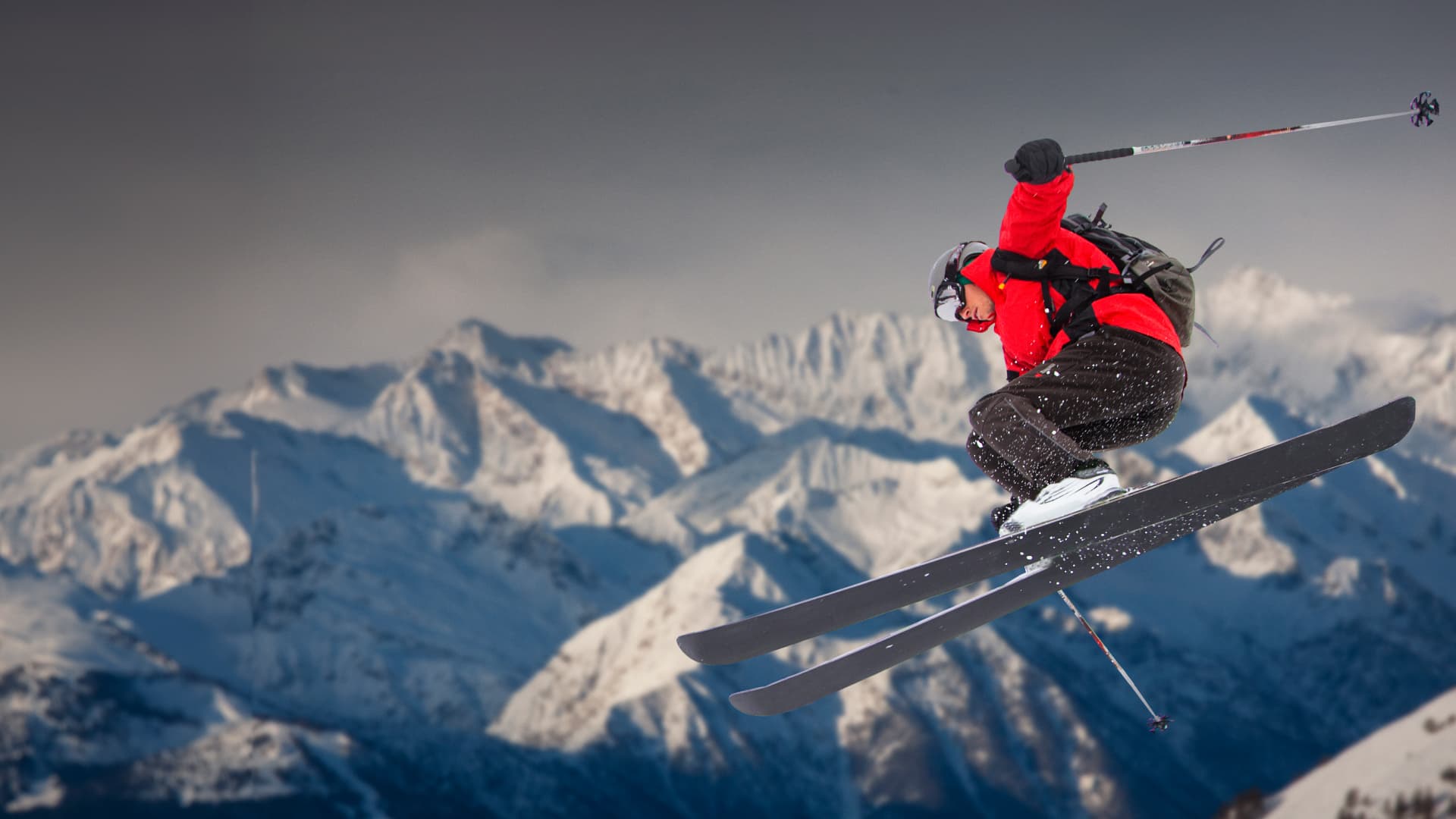 downhill skier in red jacket mid jump with snowy mountains in the background