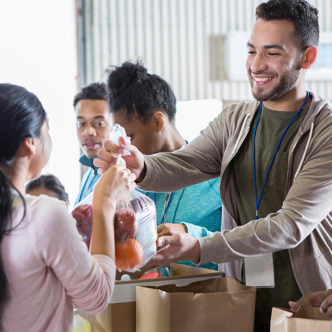 Young woman hands a bag of fruit to a young male food bank volunteer. The volunteers are receiving donations from their community to benefit a food bank.