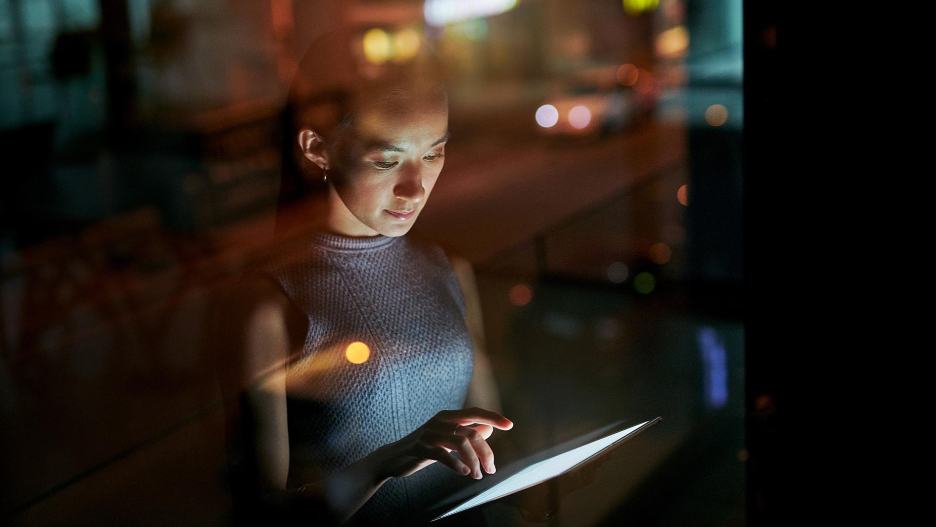 A woman standing in front of a window at night, working on ipad.