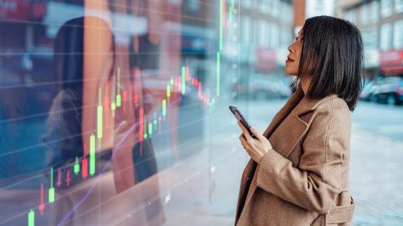Young Asian businesswoman analyzing and checking stock market over smartphone in downtown financial district. Stock exchange market trading board in background.